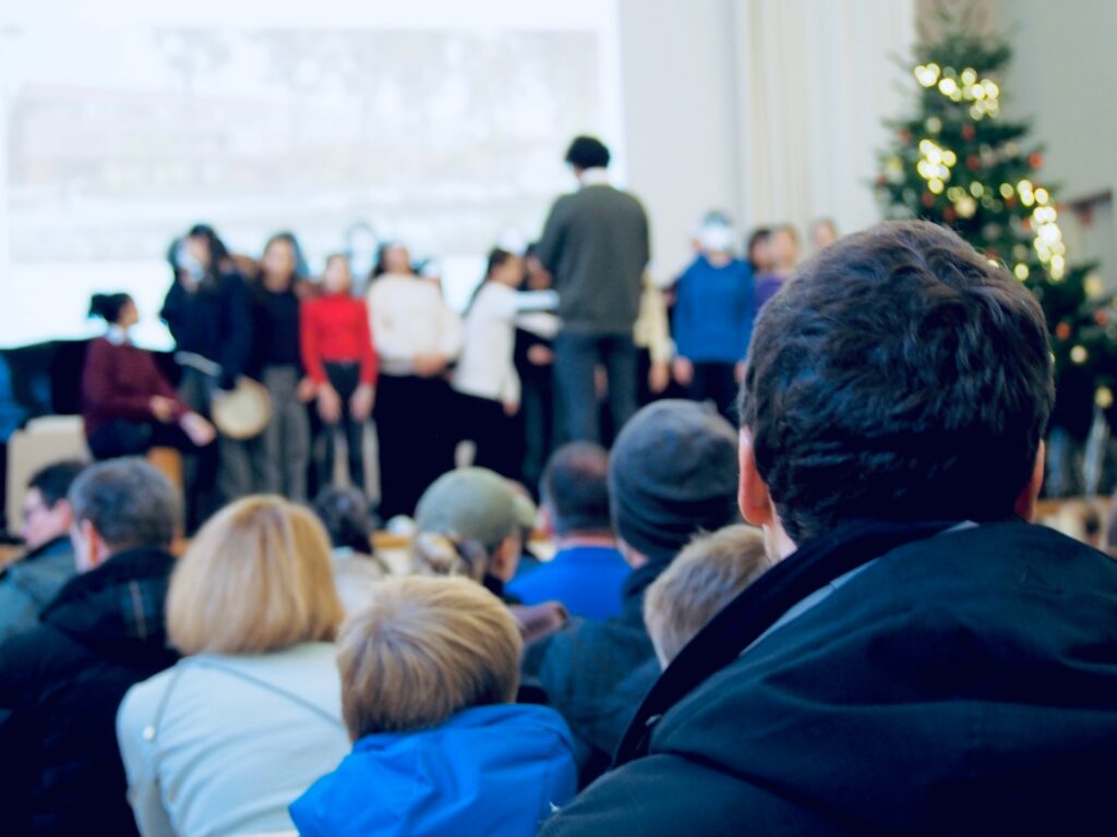 Chor auf einer Bühne vor vollem Publikum, rechts ein geschmückter Weihnachtsbaum