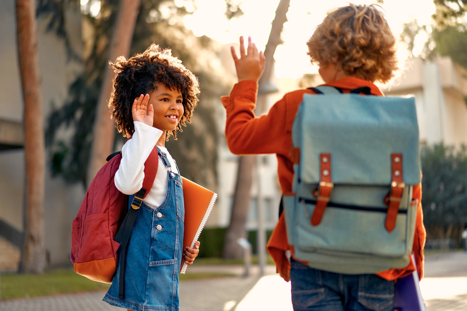 Zwei Kinder mit Schulranzen winken sich auf dem Schulweg zu